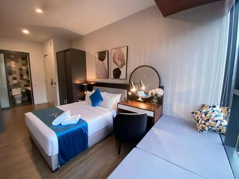 Bedroom features: queen-size bed with white bedding, upholstered headboard, wooden bedside tables, table lamps on bedside tables, dressing table with round mirror, black upholstered chair, decorative flowers in vase, wall-mounted artwork, wooden flooring, large window with city view, built-in wardrobe, grey accent wall; style: modern; view: city. Room features: toilet visible in background, grey hexagonal tile wall.