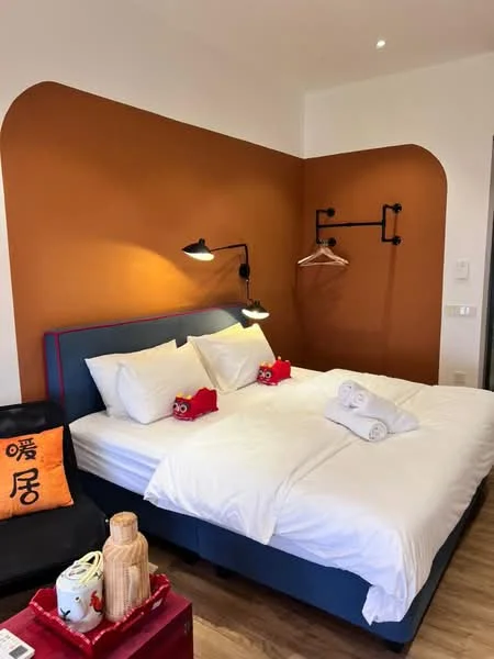Bedroom features: king-size bed, white bedding, two white towels, orange accent wall, blue upholstered headboard, black wall-mounted reading lights, wooden flooring, clothes rack, decorative red plush toys, small side table, decorative wooden container, black chair with cushion; style: modern.