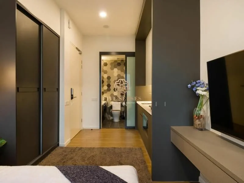 Room features: dark grey accent wall, light wood flooring, built-in wardrobe with sliding doors, wall-mounted TV, floating console table, decorative vase with flowers, recessed ceiling lights, doorway leading to bathroom, textured wallpaper in bathroom area; style: modern.