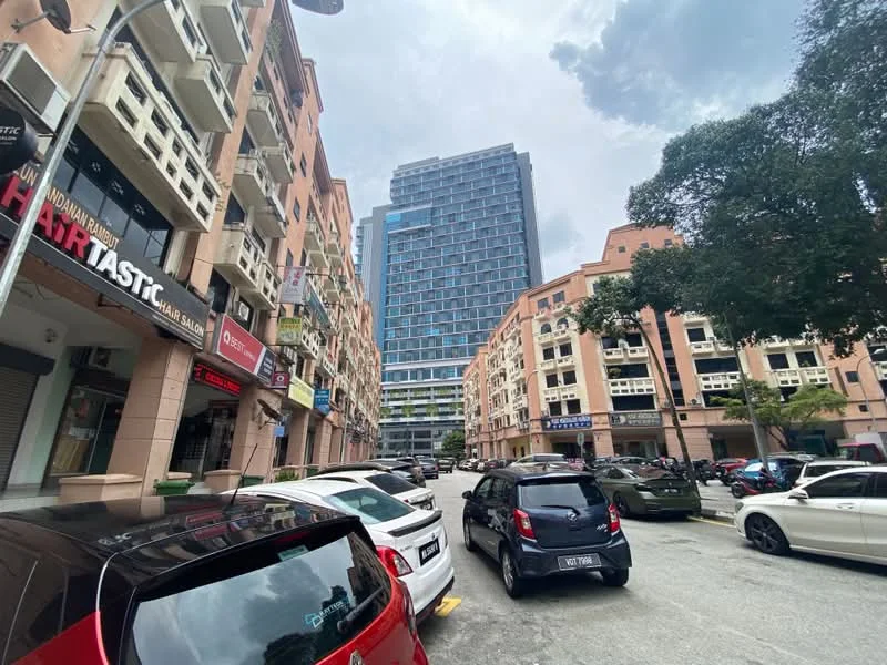 Exterior type: mixed-use development; features: multi-storey buildings, commercial units on ground floor, residential units above, glass curtain wall on high-rise, balconies on lower buildings, street view with parked cars, surrounded by trees, urban setting, visible signage on buildings, paved road; style: modern.
