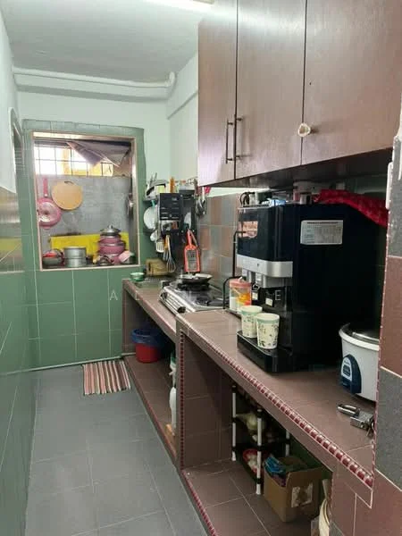 Kitchen features: green tiled walls, brown wooden cabinetry, countertop with storage underneath, stainless-steel coffee machine, rice cooker, various pots and pans, red and white striped kitchen mat, grey tiled flooring, window with metal grill, shelves with kitchen items.