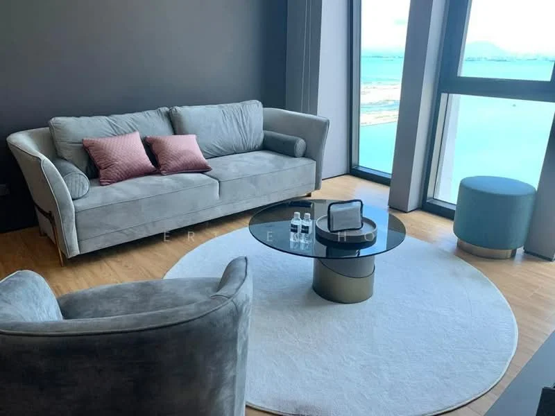 Living features: 3-seater fabric sofa in grey, 2 accent pillows in pink, round glass coffee table with gold base, area rug in circular shape, floor-to-ceiling windows, wooden flooring, view of the sea, accent chair in grey, decorative items on the coffee table; style: modern; view: sea.