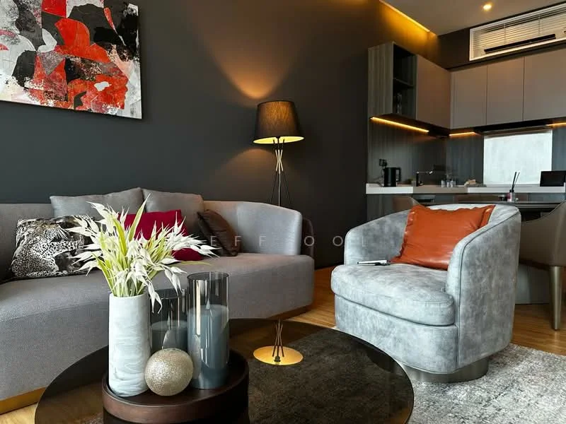 Living features: grey fabric 3-seater sofa, accent chair in light grey with orange cushion, black floor lamp, round coffee table with dark wood base, decorative vase with white flowers, glass vase, decorative sphere, abstract artwork on wall with red and black colors, recessed lighting, wooden flooring, open kitchen area visible in background; style: modern.