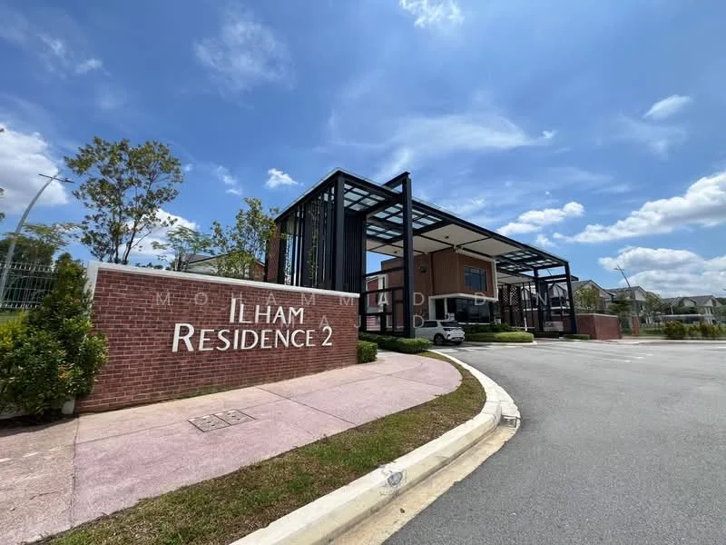 Exterior type: gated community entrance; features: modern architectural design, glass canopy, brick facade, landscaped garden, manicured shrubs, paved driveway, signage with 'ILHAM RESIDENCE 2', surrounding greenery, clear blue sky, well-maintained entrance; style: modern.