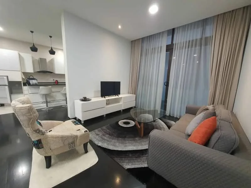 Living features: 3-seater fabric sofa in grey, accent chair with patterned upholstery, glass coffee table with wooden legs, area rug with circular pattern, wall-mounted TV, white TV console, floor-to-ceiling windows with sheer curtains, dark wood flooring, recessed downlights, open-plan layout to kitchen; style: modern.