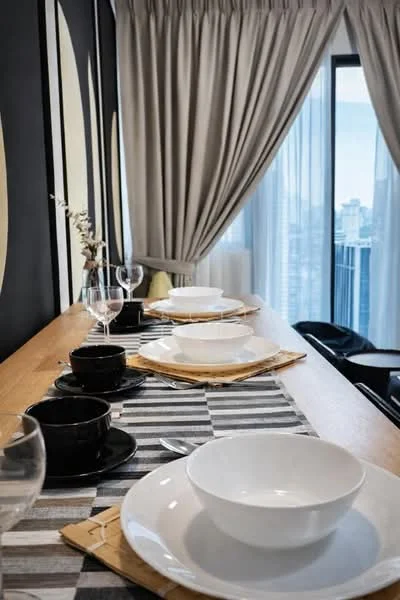 Dining features: wooden dining table, white ceramic bowls, black ceramic cups, glassware, striped table runner, grey curtains, floor-to-ceiling windows, city view; style: modern; view: city.