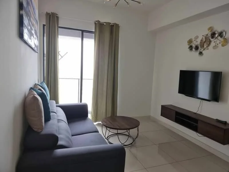 Living features: 3-seater fabric sofa in navy blue, 2 decorative cushions in light blue, round wooden coffee table with metal base, wall-mounted flat-screen TV, TV console with storage, floor-to-ceiling windows, sheer curtains in light green, light fixture with multiple bulbs, cream painted walls, tile flooring; style: modern.