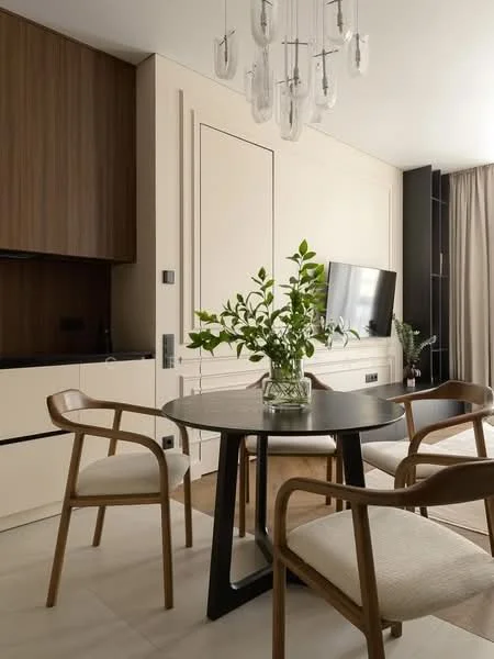 Dining features: round black dining table, 4 wooden dining chairs with fabric seats, chandelier with glass bulbs, cream painted walls, wooden cabinetry in dark brown, built-in wall unit with TV, floor-to-ceiling curtains, light-colored tile flooring, decorative plant in glass vase; style: modern.