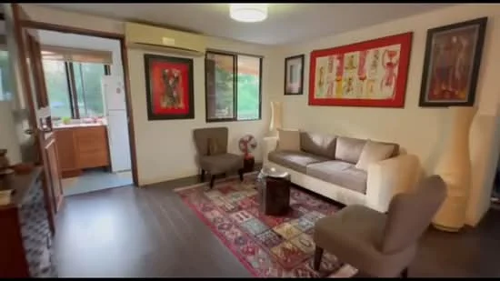 Living features: 3-seater fabric sofa in light grey, 2 upholstered armchairs in grey, wooden coffee table, area rug with geometric pattern, wall-mounted artwork in frames, floor-to-ceiling windows, air conditioning unit, laminate flooring, decorative floor vases, light ceiling fixture; style: contemporary; view: garden. Kitchen features: wood cabinetry, countertop, kitchen appliances visible, open layout to living area.