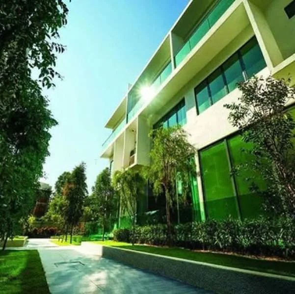 Exterior type: low-rise apartment block; features: glass curtain wall, green tinted windows, white painted facade, manicured lawn, landscaped garden with shrubs, stone-paved walkway, surrounded by mature greenery; style: modern.