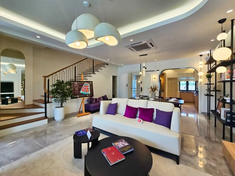 Living features: cream painted walls, marble tile flooring, tray ceiling, 3-blade ceiling fan, pendant lamps, sofa with cream upholstery, purple accent cushions, coffee table, decorative plants in ceramic pots, wall-mounted TV, open shelving unit, staircase with wooden railing, area rug with geometric pattern; style: modern.