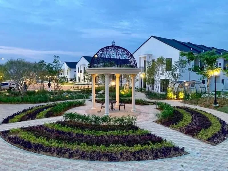 Exterior type: landscape garden; features: decorative gazebo with dome roof, paved walkway, manicured flower beds, landscaped garden with shrubs, outdoor seating area with chairs, ambient lighting around garden, surrounding residential buildings, greenery and trees in background; style: modern.