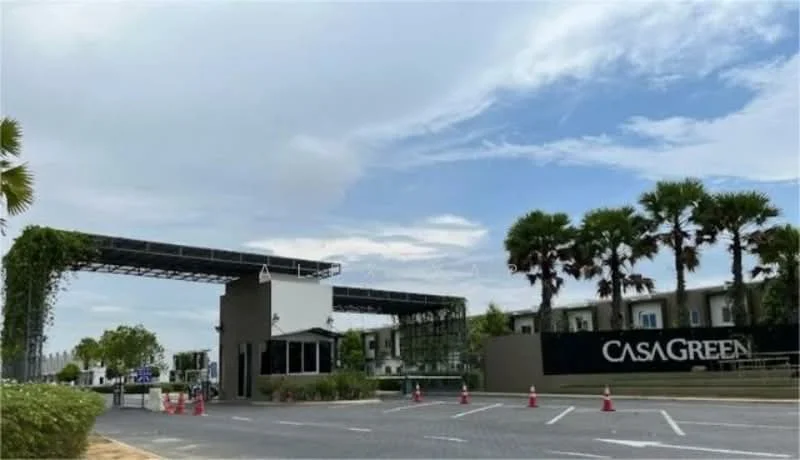 Exterior type: gated community; features: main entrance gate, guard house at entrance, landscaped greenery, palm trees, paved driveway, parking lot, signage with 'CASA GREEN', surrounded by low-rise buildings, open-air parking, security cones; style: modern.