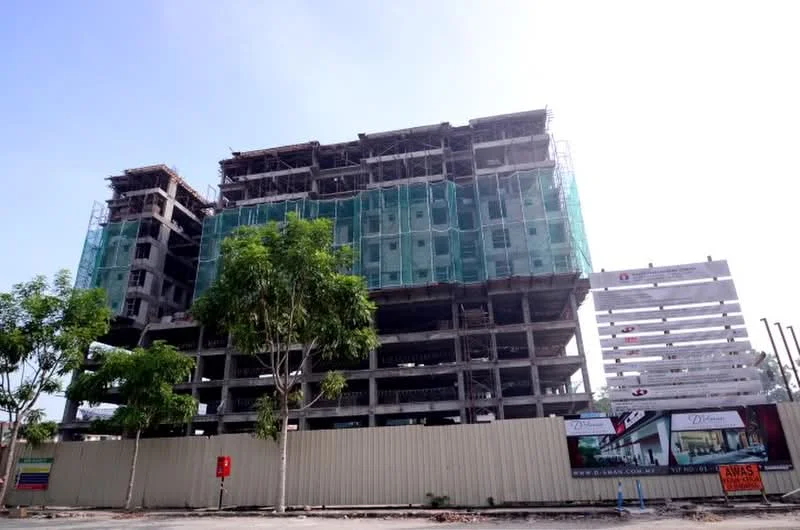 Exterior type: mid-rise condominium; features: unfinished concrete facade, scaffolding visible, green construction netting, multiple storeys, surrounding trees, construction hoarding, advertisement signage, open-air parking area; style: modern.