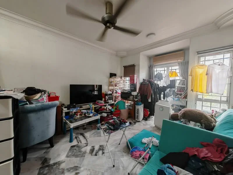 Living features: 5-blade ceiling fan, wall-mounted TV, light blue fabric sofa, marble tile flooring, white painted walls, large window with sheer curtains, clothes drying rack with clothes, various storage boxes, coffee table, decorative items on shelves, messy arrangement of items, open space layout.