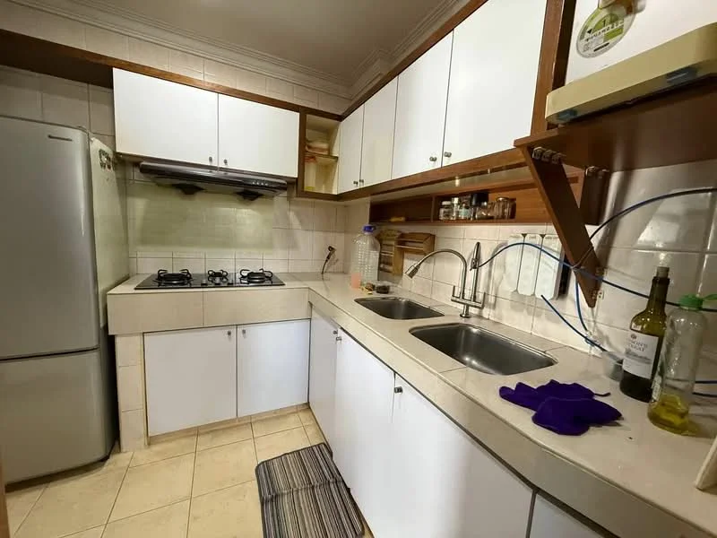Kitchen features: white cabinetry, stainless-steel fridge, built-in gas hob with 2 burners, double-bowl stainless-steel sink, chrome pull-down faucet, cream tiled backsplash, light-colored countertop, tile flooring, wall-mounted range hood, kitchen towel on countertop, bottle of wine on countertop, plastic water bottle on countertop.