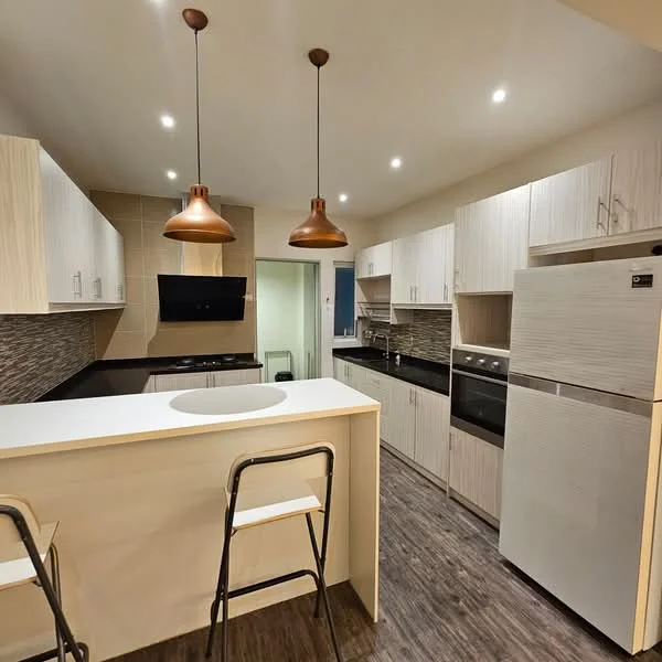 Kitchen features: light wood cabinetry, black countertop, stainless-steel fridge, built-in oven, 4-burner gas hob, black backsplash with tile pattern, single-bowl stainless-steel sink, chrome pull-down faucet, 2 pendant lights, bar counter with seating, wood laminate flooring, recessed LED downlights; style: modern.