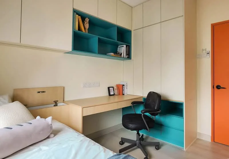 Bedroom features: single bed with white bedding, built-in desk with chair, open shelving unit with teal backing, cream-colored cabinetry, orange door, light-colored flooring, decorative items on shelves, wall-mounted electrical outlets; style: modern.