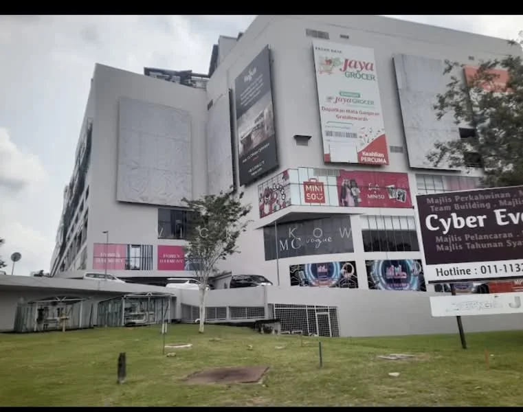 Exterior type: shopping mall; features: modern facade with large glass windows, advertisement billboards on exterior walls, green landscaping with grass area, multiple levels visible, entrance with covered walkway, parking area visible in front, signage for various shops, surrounded by trees; style: modern.