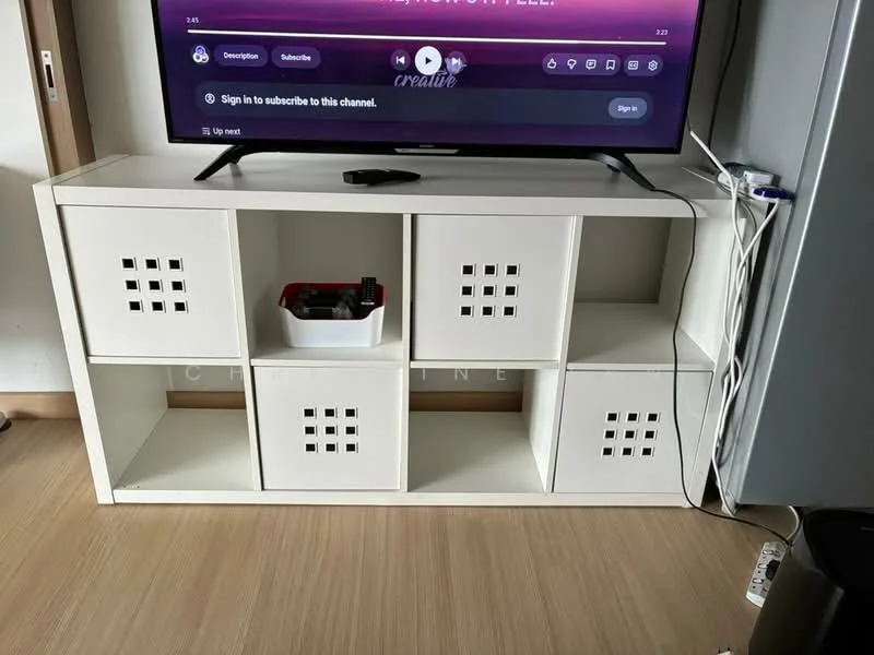 Room features: white shelving unit with square cutouts, open shelving design, light wood flooring, TV console with electronic devices, cable management visible, black trash bin, decorative items on shelves; style: modern.