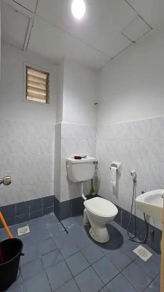 Bathroom features: white tiled walls, light grey tiled flooring, one-piece toilet, wall-mounted toilet tank, sink with faucet, toilet paper holder, window with horizontal slats, shower area with drainage, cleaning bucket, wall-mounted mirror.