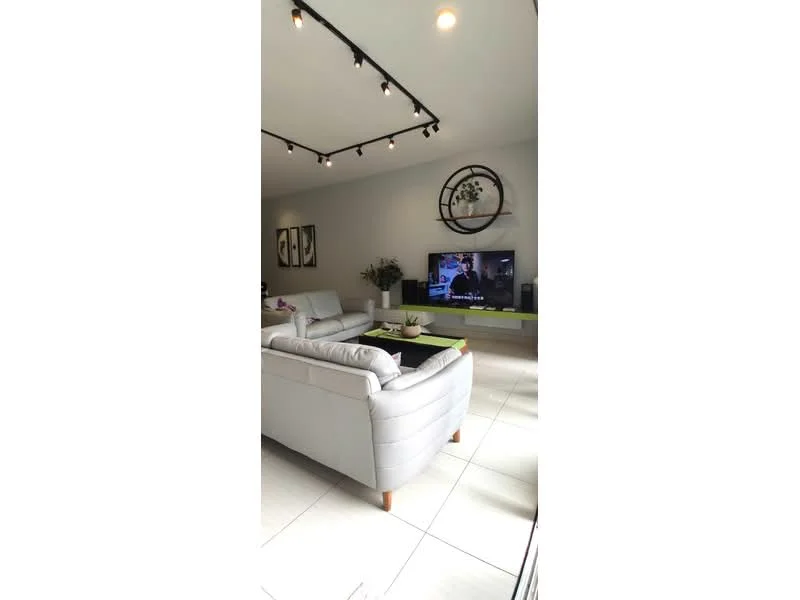 Living features: light grey leather sofa, 2-seater sofa, coffee table, TV console, wall-mounted TV, recessed LED downlights, decorative wall clock, framed artwork on walls, green accent table, floor tiles in light color, plant in decorative pot; style: modern.