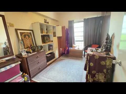 Room features: wooden flooring, cream painted walls, large window with grid pattern, dark curtains, brown wooden dresser, decorative wall art, small storage cabinet, orange chair, decorative table with cloth, statues and figurines on display, area rug with geometric pattern; style: traditional; view: garden.