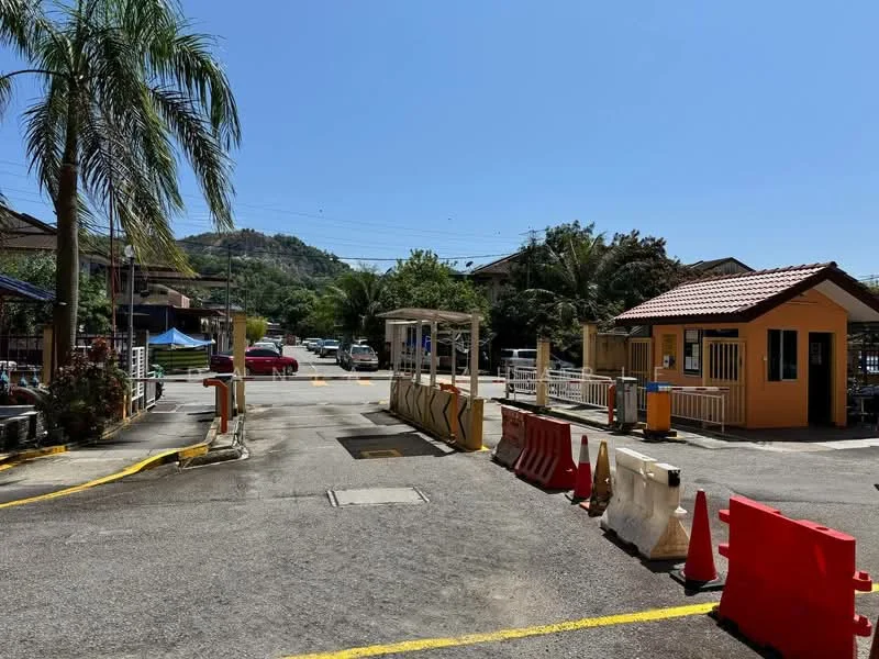 Exterior type: main entrance gate; features: guard house at entrance, palm trees, barrier cones, yellow road markings, surrounding greenery, residential area in background, clear blue sky, paved road; style: modern.