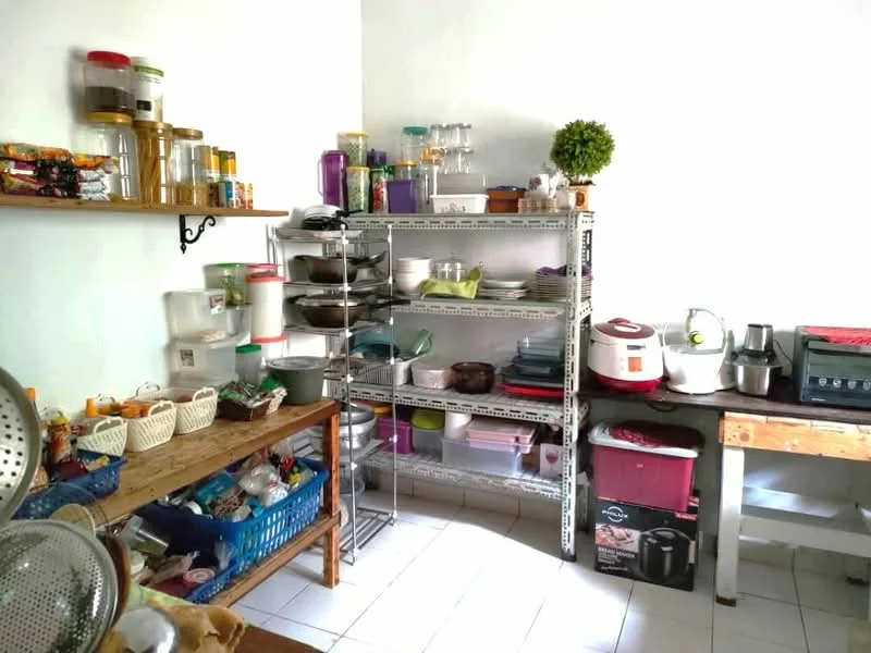 Kitchen features: white painted walls, white tiled flooring, metal shelving unit with various containers, wooden table, blue plastic basket, rice cooker, blender, stainless-steel colander, decorative plant, multiple jars and bottles on shelves, cooking pots and pans, microwave, food storage containers.