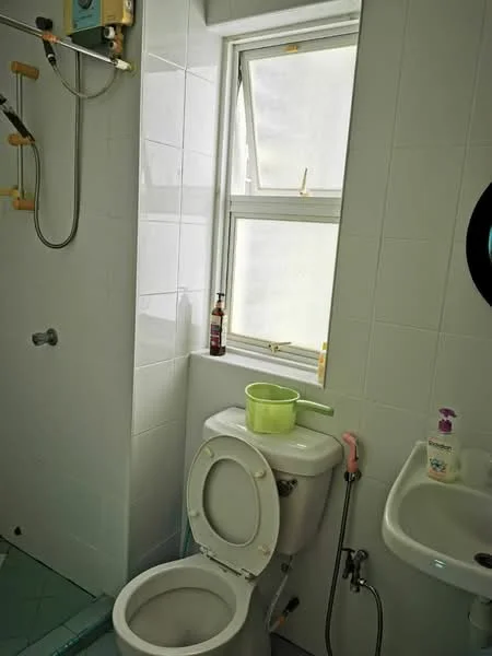 Bathroom features: white tiled walls, white tiled flooring, one-piece toilet, sink with chrome faucet, shower with wall-mounted showerhead, window with white frame, green plastic bucket, soap dispenser, bottle on windowsill.