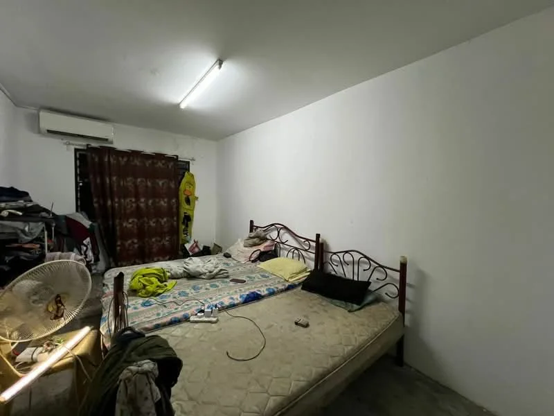 Bedroom features: double bed with metal frame, mattress with bedding, wall-mounted air conditioning unit, white painted walls, concrete flooring, curtains, fan, clothes piled on the floor, bedside items on the bed.