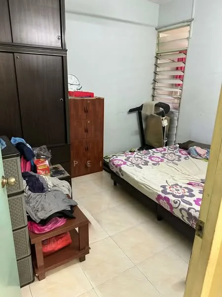 Bedroom features: queen-size bed with floral bedding, wooden wardrobe with sliding doors, wooden chest of drawers, exercise equipment, tile flooring, white painted walls, window with horizontal slats, small bedside table, clothes on the floor.