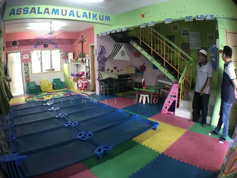 Room features: colorful rubber flooring, green painted walls, pink painted walls, decorative wall with 'ASSALAMUALAIKUM' text, decorative wall with 'WELCOME' text, staircase with railing, play area with mats, seating area with chairs, decorative wall art, children's play equipment, large circular rug, storage shelves, window with curtains; style: playful; view: interior.