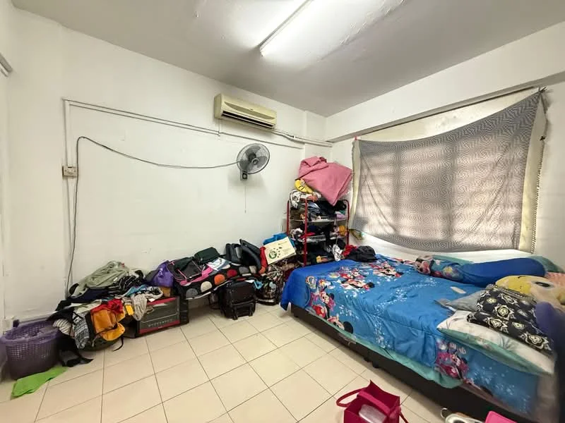 Bedroom features: single bed with colorful bedding, wall-mounted air conditioning unit, ceiling light fixture, white painted walls, tiled flooring, bookshelf with various items, cluttered with bags and personal items, window with curtain, fan on the wall.