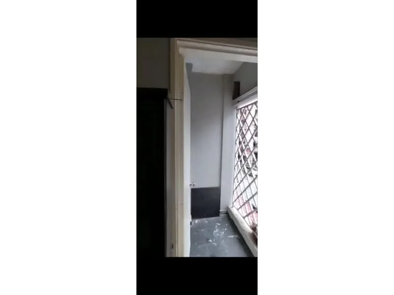 Room features: grey painted walls, black tiled flooring, window with metal grille, light fixture, corner space, unfinished wall, visible debris on floor.