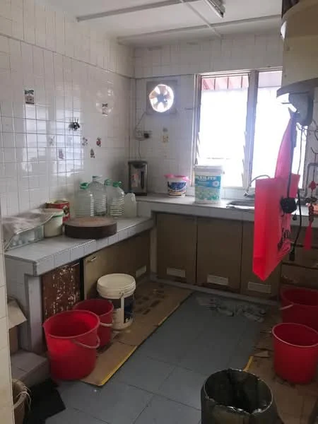 Kitchen features: white tiled walls, grey tiled flooring, window with sliding shutters, single-bowl stainless-steel sink, chrome faucet, wooden cabinetry, countertop with storage underneath, multiple plastic buckets, water dispenser, circular cutting board, wall-mounted ventilation fan.