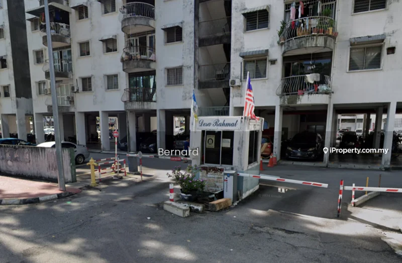 Exterior type: low-rise apartment block; features: main entrance gate, guard house at entrance, covered parking, multiple storeys, balconies on upper floors, visible vehicles in parking area, surrounded by concrete pavement, security barrier at entrance; style: modern.