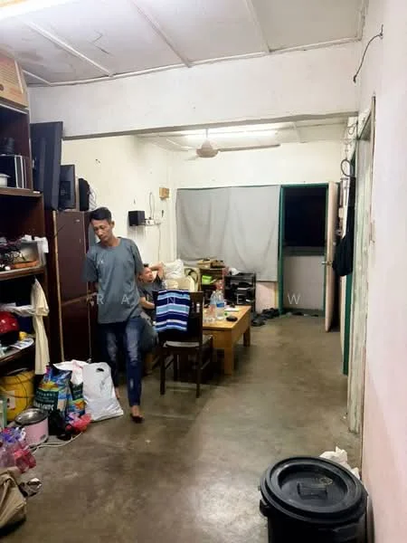 Room features: concrete flooring, white painted walls, ceiling fan, dining table with chairs, wooden shelving unit, television on shelf, curtains on window, plastic storage bins, various bags and items on floor, doorway leading to outside.