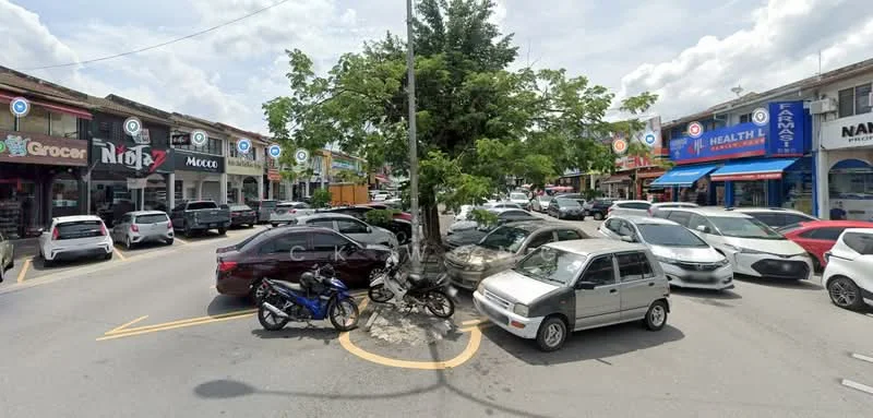 Exterior type: shophouse; features: street view with multiple parked cars, tree in the center of the roundabout, various commercial shopfronts, signage visible on buildings, pedestrian pathways, multiple storefronts with awnings, urban setting with cloudy sky; style: modern.