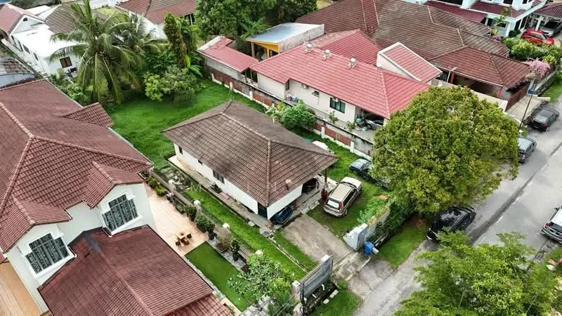 Exterior type: detached bungalow; features: red tiled roof, manicured lawn, palm trees, surrounded by greenery, driveway with interlocking pavers, single-storey, adjacent houses with similar roofing, garden area, car parked in driveway, fenced perimeter; style: traditional.