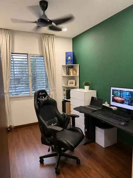 Study features: green painted walls, wood laminate flooring, ceiling fan, window with horizontal blinds, desk with computer monitor, black office chair, white storage cabinets, bookshelf with decorative items, potted plant; style: modern.
