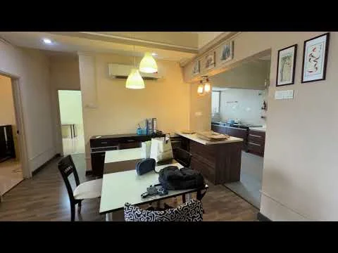 Dining features: dining table with white surface, 4 upholstered dining chairs, pendant lighting above dining table, wooden flooring, wall-mounted air conditioning unit, decorative wall art, open layout to kitchen, dark wood cabinetry; style: modern. Kitchen features: dark wood cabinets, countertop with integrated sink, stainless-steel appliances, open layout to dining area, kitchen island, recessed lighting; style: modern. Room features: light-colored walls, doorway leading to another room.
