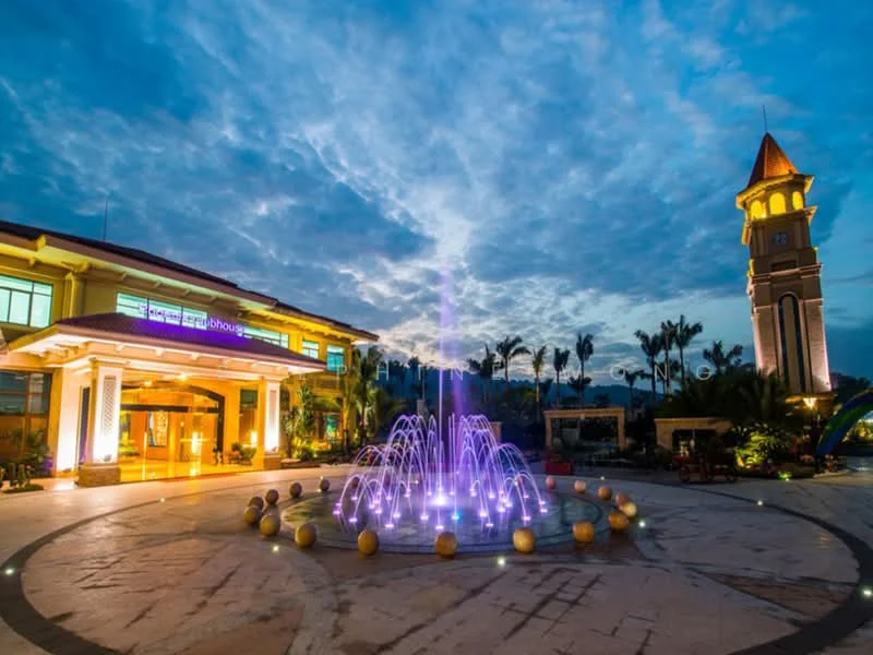 Exterior type: clubhouse; features: fountain with colored lights, landscaped garden, paved walkway, clock tower, illuminated building facade, outdoor seating area, mature palm trees, night sky with clouds; style: modern.