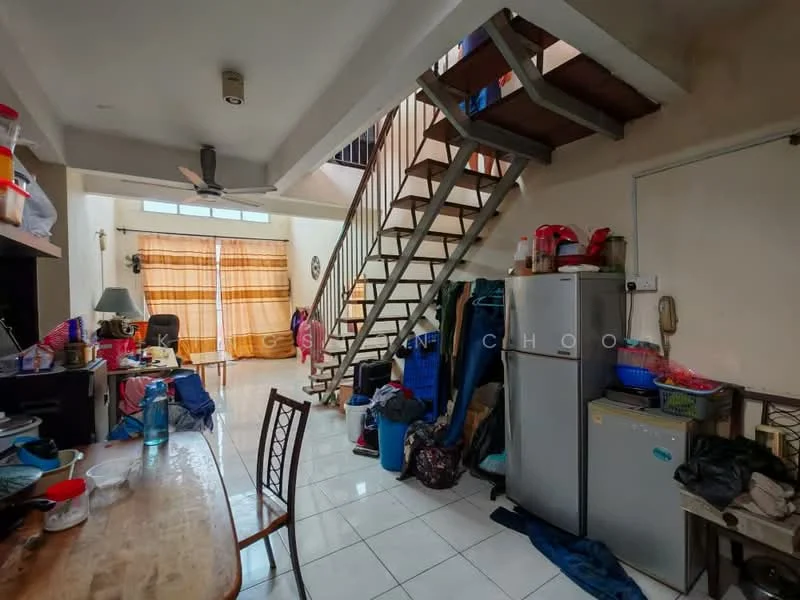 Room features: white painted walls, ceramic tile flooring, exposed staircase, ceiling fan, dining table, wooden chair, stainless-steel fridge, plastic storage containers, blue tarpaulin, various bags and items on the floor, window with sheer curtains, kitchen area with clutter.