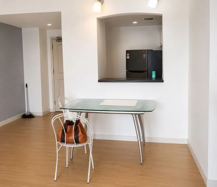 Dining features: glass dining table, white metal chairs, light wood flooring, grey accent wall, refrigerator, open kitchen pass-through, two ceiling lights, neutral painted walls; style: modern.