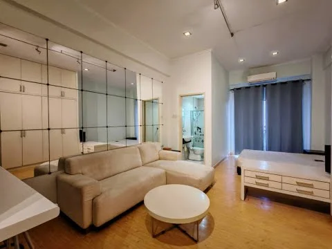 Living features: beige fabric L-shape sectional sofa, round white coffee table, built-in wardrobe with mirrored doors, wooden flooring, recessed downlights, floor-to-ceiling windows with sheer curtains, wall-mounted air conditioning unit, light-colored walls; style: modern. Room features: bathroom visible through door, white walls, light-colored flooring.