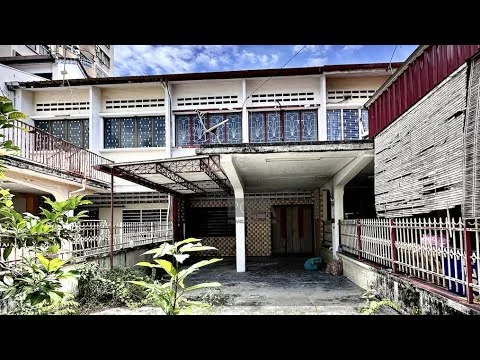 Exterior type: linked terrace house; features: two-storey building, red painted facade, balcony with railing, exposed concrete structure, multiple windows with grilles, front yard with plants, covered car porch, brick wall fencing, visible basketball hoop, tile flooring at entrance, manicured garden area, surrounded by residential buildings; style: traditional.