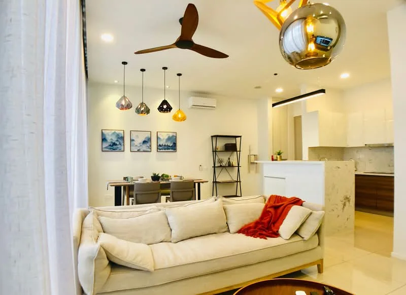 Living features: 3-seater fabric sofa in cream, red throw blanket on sofa, round coffee table, 5-blade ceiling fan with light, pendant lights in various colors, wall-mounted air conditioning unit, marble countertop on kitchen island, open-plan layout to dining area, light-colored walls, tile flooring, decorative wall art with landscape prints, black shelving unit; style: modern. Dining features: 4-seater dining table, grey dining chairs, table setting with plates and cups, light fixture above dining table; style: modern.