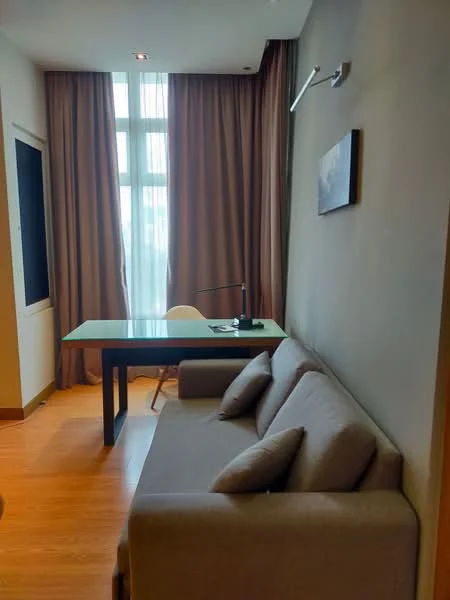 Study features: brown painted walls, wooden flooring, floor-to-ceiling windows, sheer curtains, desk with glass top, white chair, sofa in grey fabric, table lamp, framed artwork on wall; style: modern.