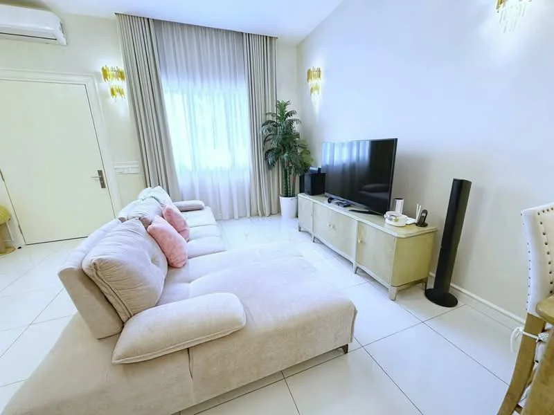 Living features: L-shape sectional sofa in light beige, 2 decorative pillows in pink, flat-screen TV on a light wood TV console, floor lamp in black, large window with sheer curtains, potted plant in the corner, white tiled flooring, wall-mounted air conditioning unit, gold wall sconces; style: modern.